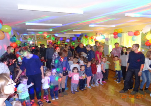 Dzieci i dorośli stoją na sali udekorowanej balonami w oczekiwaniu na kolejną zabawę. Na sali są kolorowe światła.
